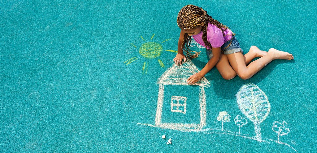 Imagem mostra criança desenhando com giz uma casa e um sol no chão azul