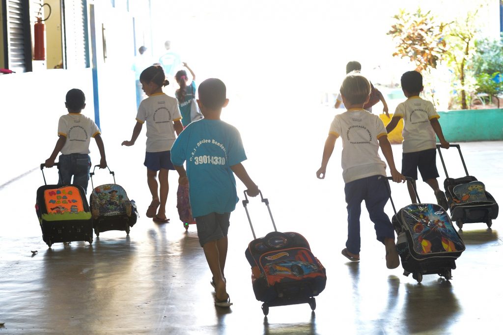 crianças de costas indo para a escola arrastando suas mochilas com rodinhas